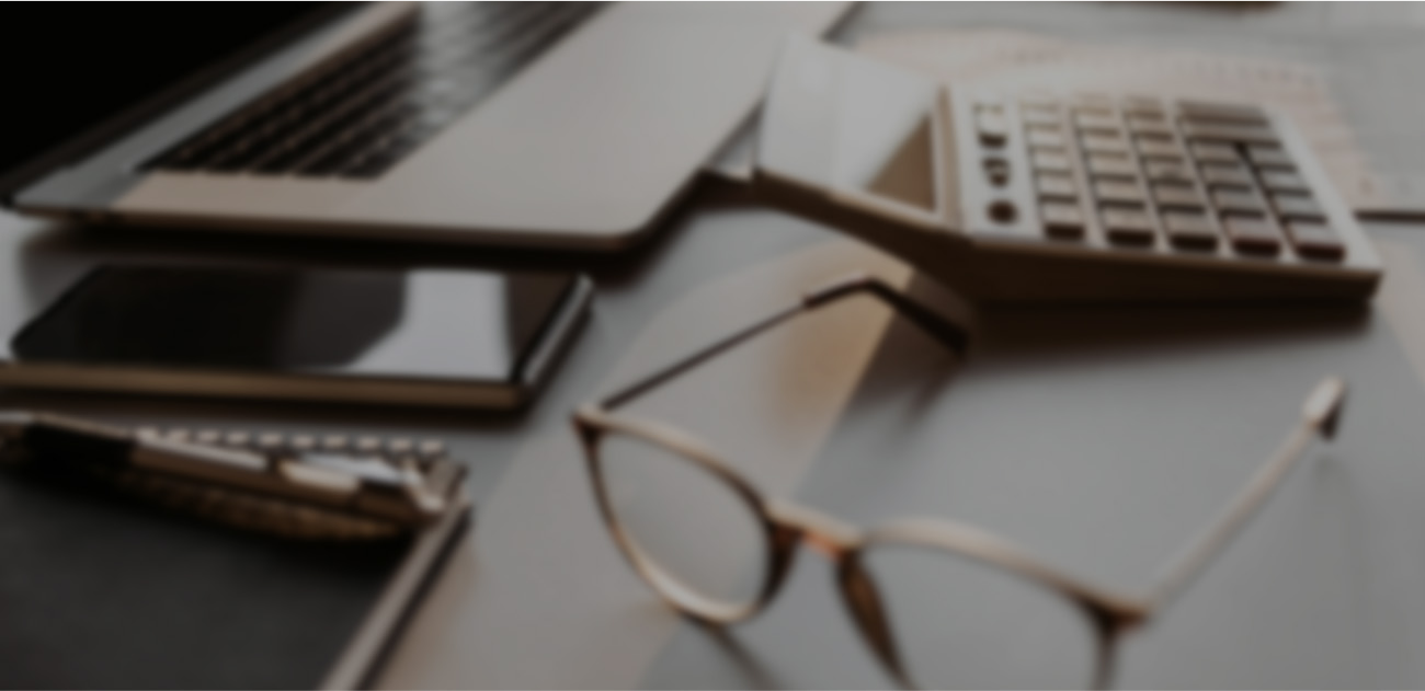 Blurred image of a workspace with a laptop, smartphone, calculator, pen, and eyeglasses on a desk, conveying a busy, professional atmosphere.