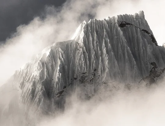 Black and white photo of a jagged snow-covered mountain peak emerging from dense clouds.