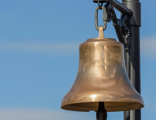 Brass bell hanging from a black iron post against a blue sky.