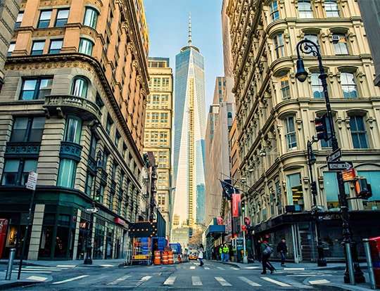 Street-level view of Lower Manhattan looking toward One World Trade Center, flanked by historic Beaux-Arts office buildings